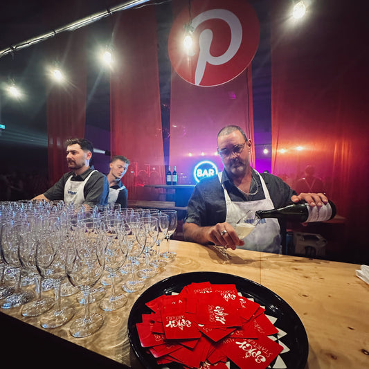 Two bartenders behind a bar with glasses and a tray of red cards, with a Pinterest logo in the background.